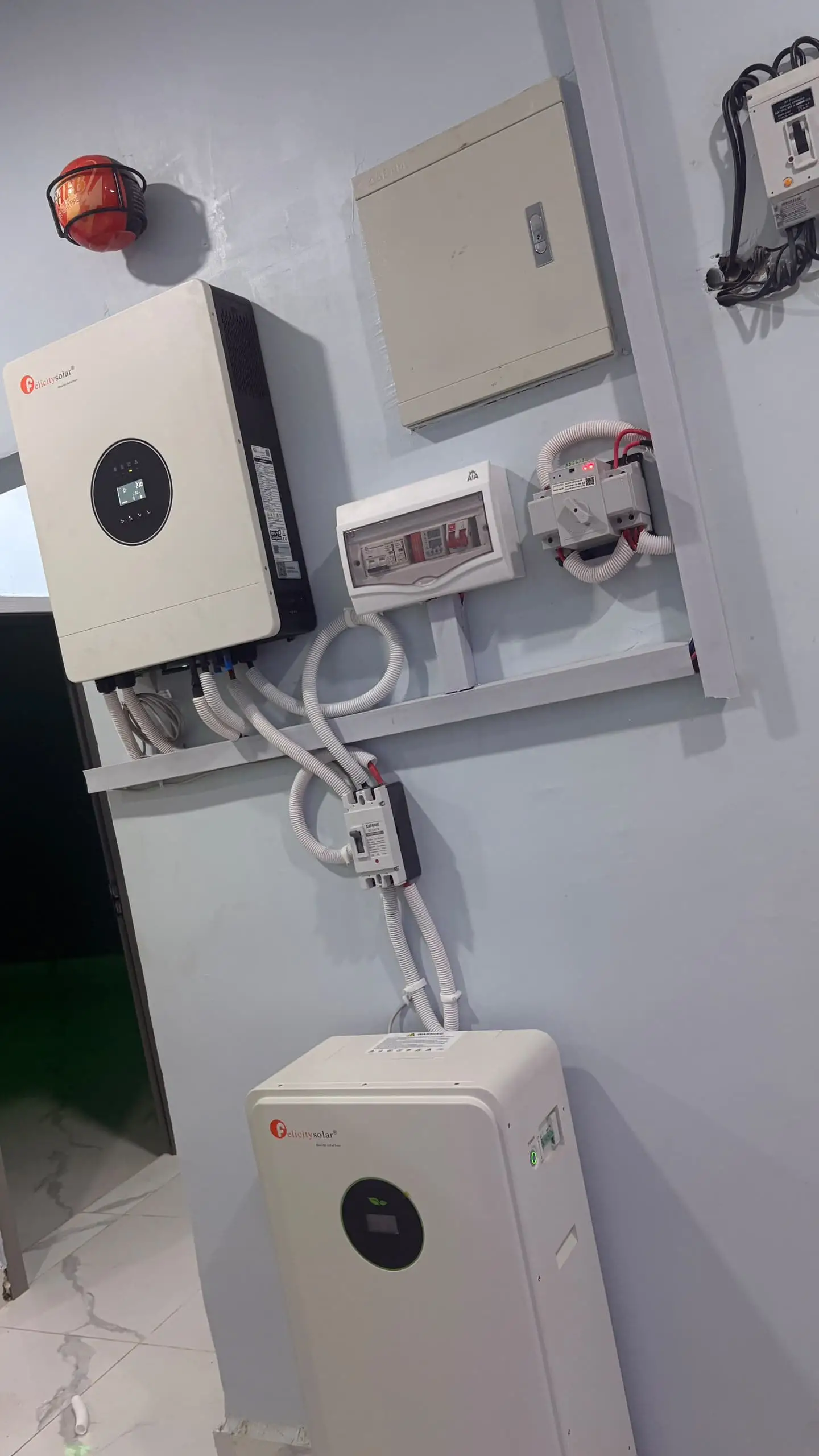 A Felicity hybrid inverter mounted on the wall beside a distribution box and changeover switch, conduit curving down to a floor-standing lithium battery below.