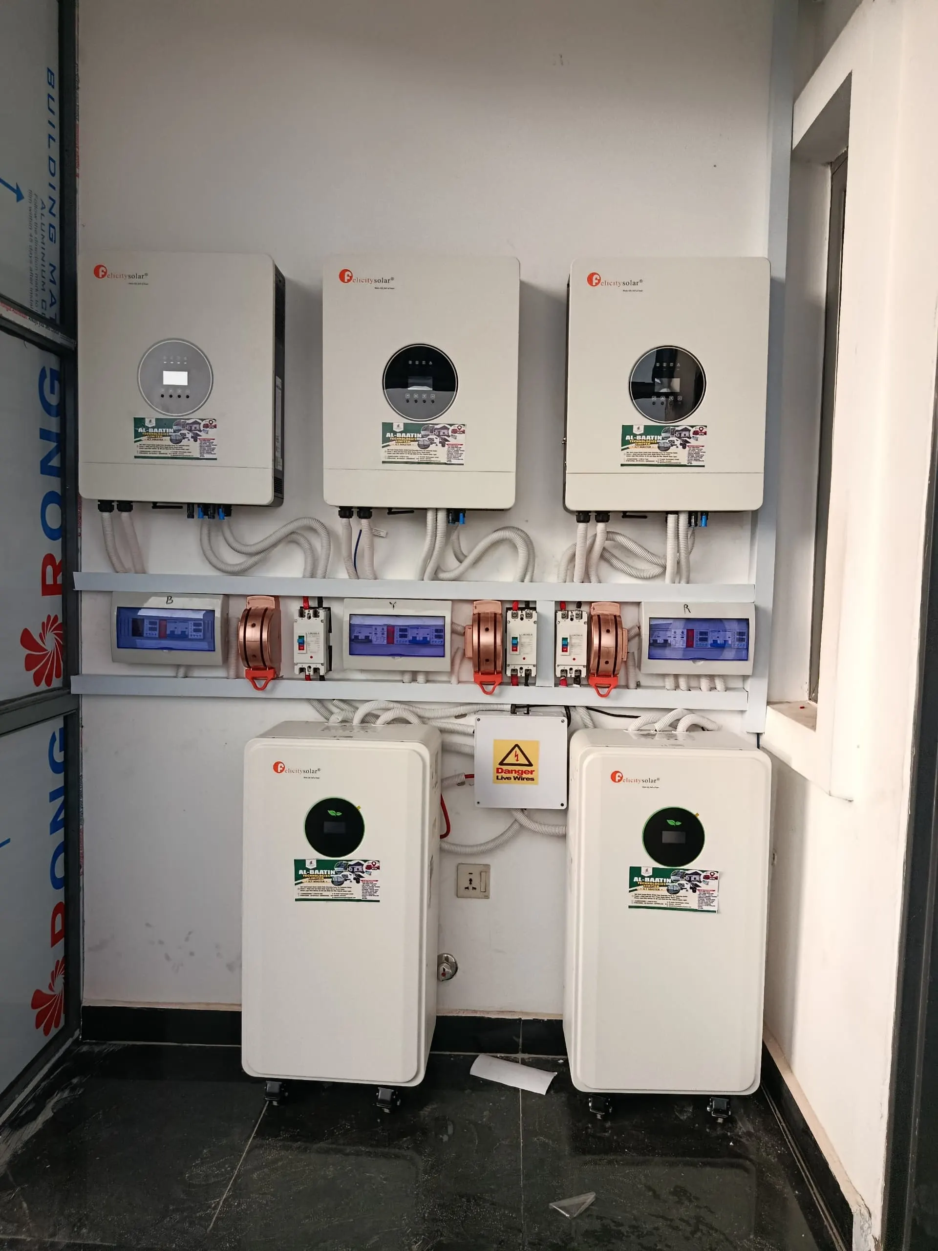 Three Felicity hybrid inverters mounted in a row above a rail of breakers and isolators, with two floor-standing lithium batteries below and a 'Danger live wires' label between them.